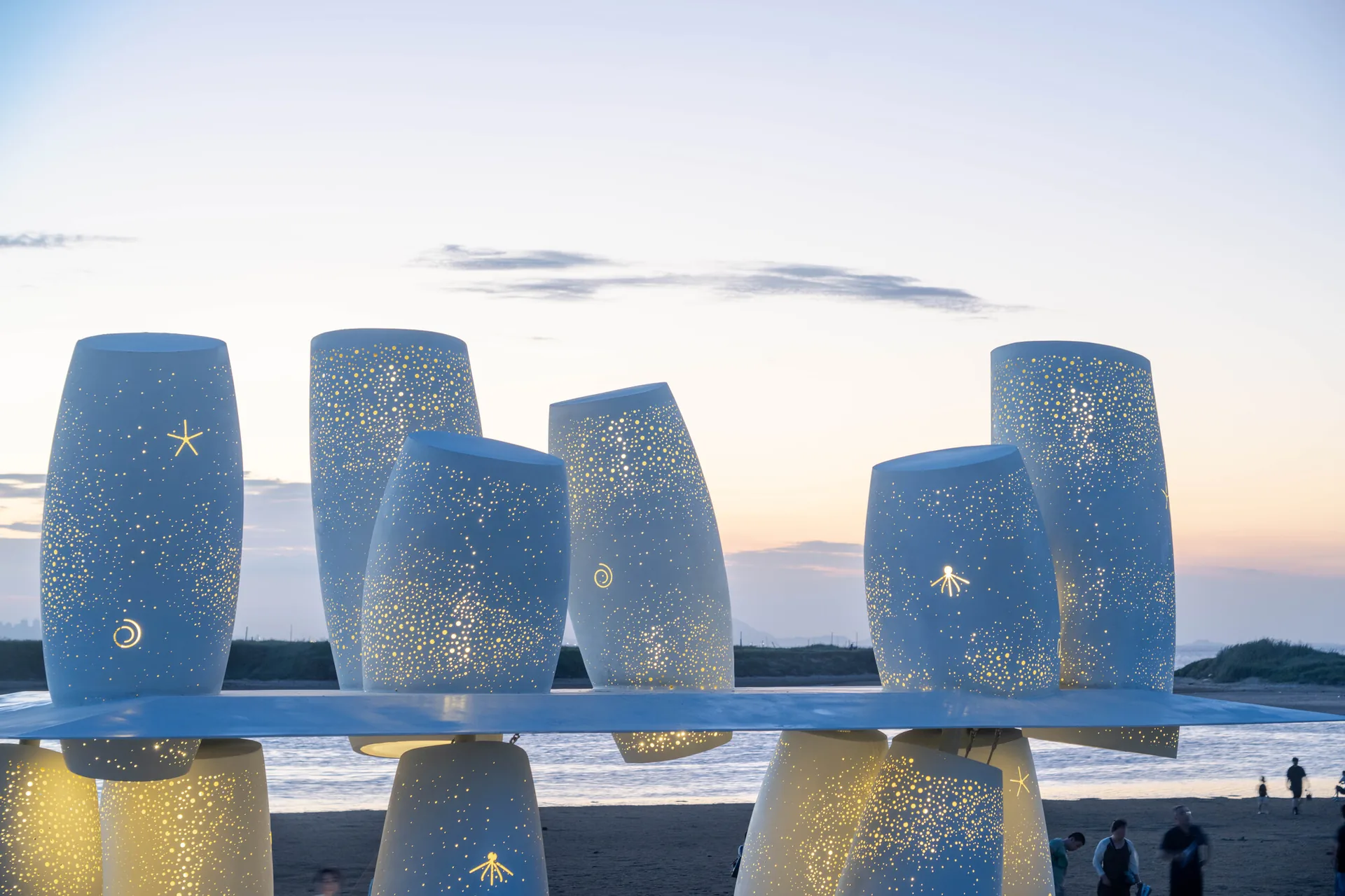 Upper cylinders lit from inside so the perforations shine like points of light after sunset, Seaside Swing Installation by XBTW OFFICE
