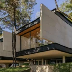 Cantilevered concrete volumes on dark steel bands rising among tall trees on a grassy slope