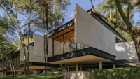 Cantilevered concrete volumes on dark steel bands rising among tall trees on a grassy slope