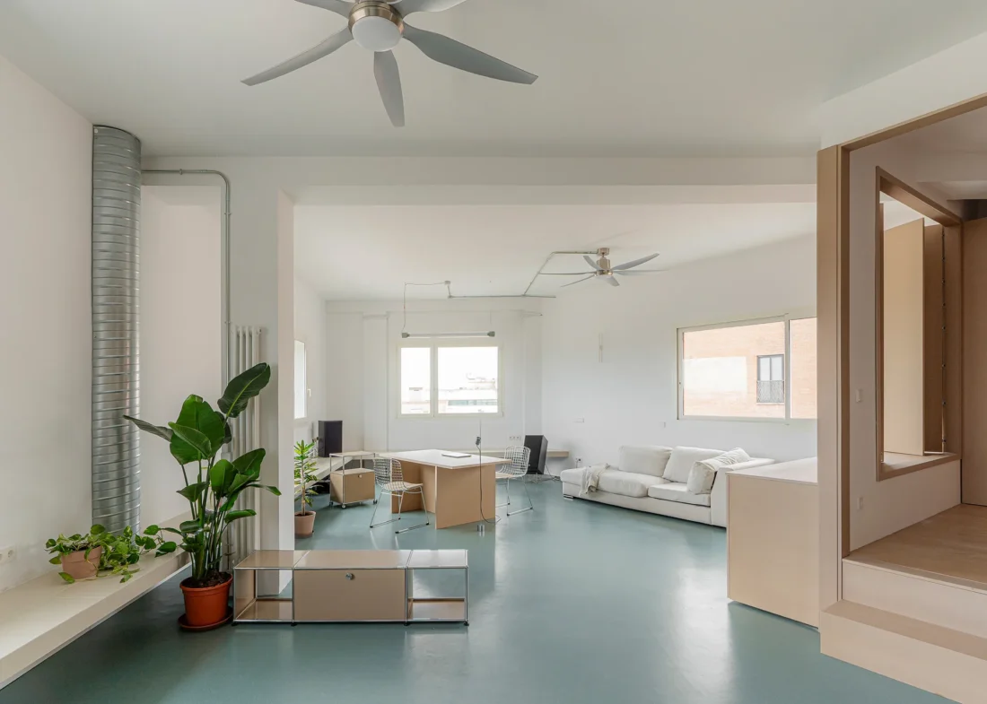 Open plan interior with a ceiling fan, plants, a white sofa and a raised wooden alcove on a blue green floor, Aguacate Space by ALE Estudio