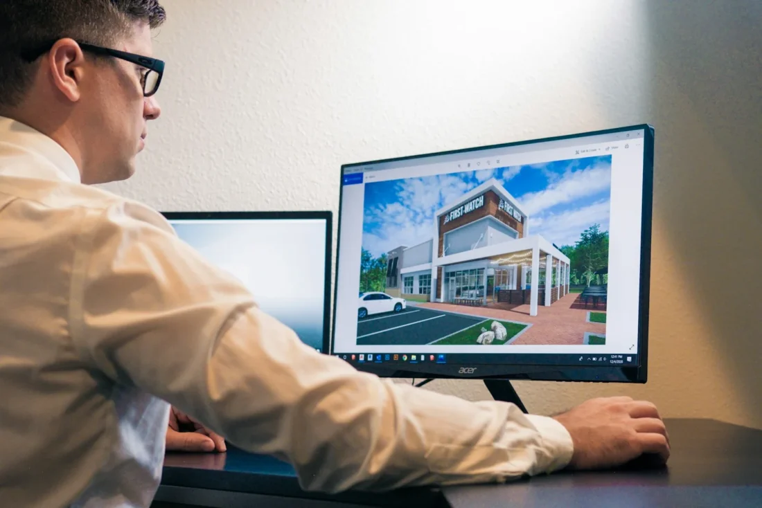Man in glasses at a desk looking at a monitor showing a rendering of a modern building