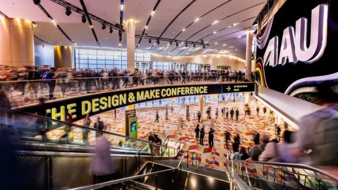 Busy convention hall entrance with escalators, a patterned carpet and a large yellow sign reading The Design and Make Conference