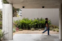 Person walking past a concrete column through a covered space with tropical plants beyond