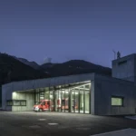 Fire station at dusk with its glazed vehicle bay lit up and a concrete hose tower carrying the brigade emblem, Civil Protection Centre by Roland Baldi Architects