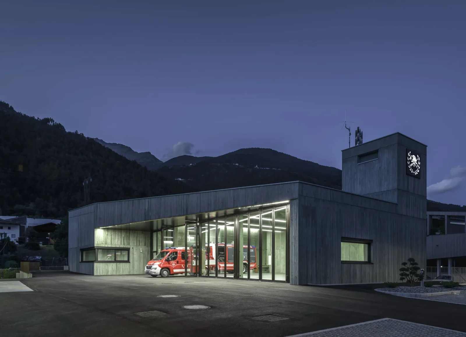 Fire station at dusk with its glazed vehicle bay lit up and a concrete hose tower carrying the brigade emblem, Civil Protection Centre by Roland Baldi Architects