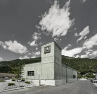 Green concrete hose tower with the fire brigade emblem rising above a low monolithic volume against wooded slopes, Civil Protection Centre by Roland Baldi Architects