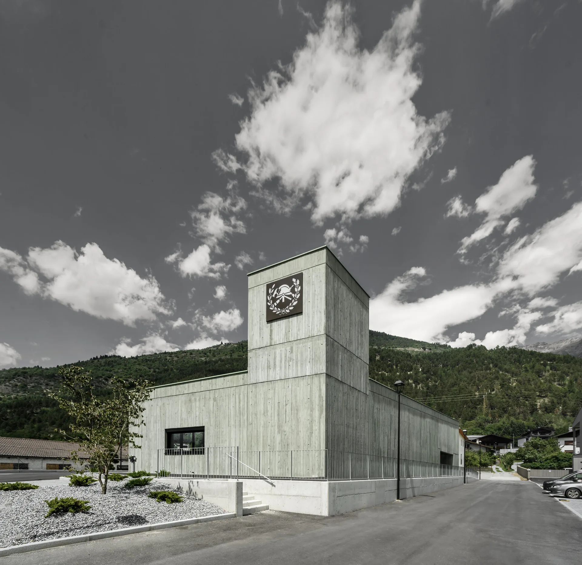 Green concrete hose tower with the fire brigade emblem rising above a low monolithic volume against wooded slopes, Civil Protection Centre by Roland Baldi Architects