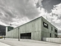 Corner of the green insulating concrete building with deep set windows and a recessed garage door along the street, Civil Protection Centre by Roland Baldi Architects