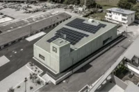 Aerial view of the trapezoidal building with solar panels on the roof, the tower and the surrounding village, Civil Protection Centre by Roland Baldi Architects