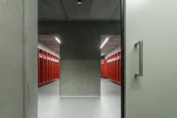 Changing room with red lockers seen through a green concrete doorway and a steel door handle, Civil Protection Centre by Roland Baldi Architects