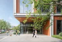 Ground floor entrance recessed under a bronze soffit with people crossing the paved forecourt, Greentown ThinkWorks