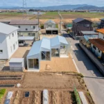 Aerial view of a row of small white gabled volumes beside a street, with vegetable plots in front and farmland beyond, House OY by 1-1 Architects