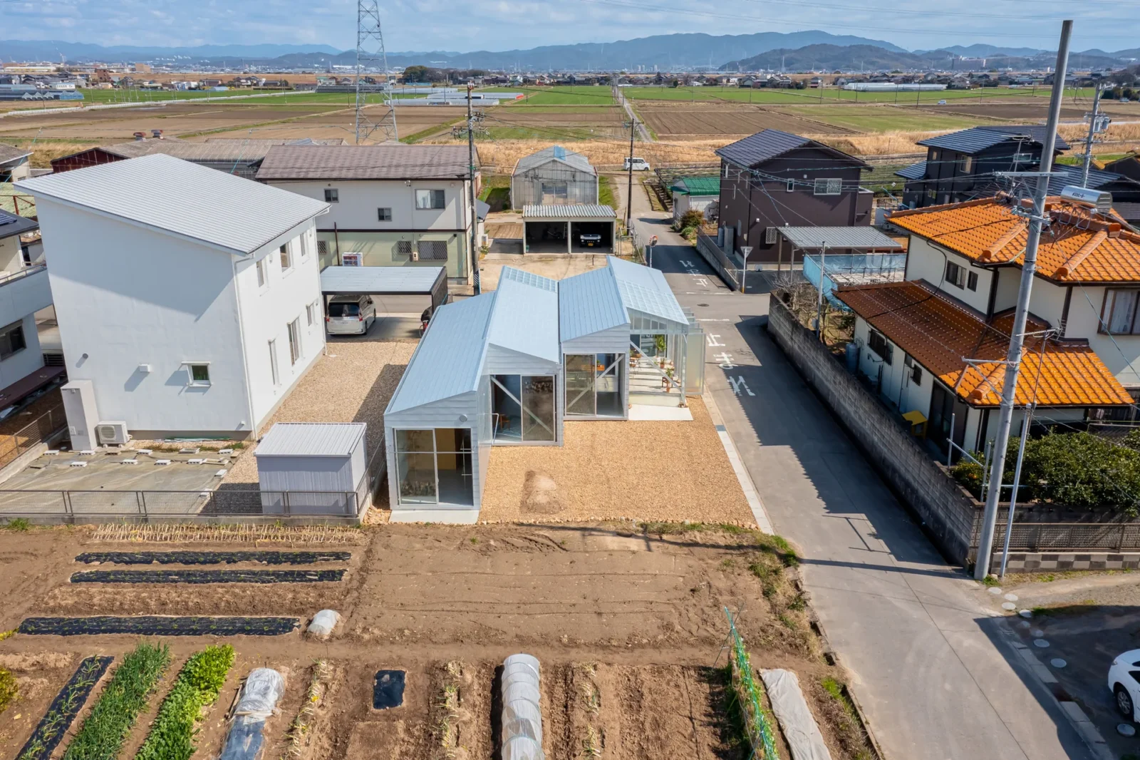Aerial view of a row of small white gabled volumes beside a street, with vegetable plots in front and farmland beyond, House OY by 1-1 Architects