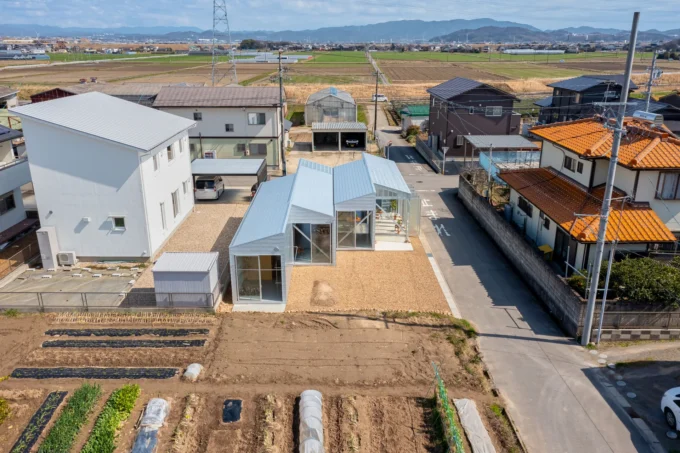 Aerial view of a row of small white gabled volumes beside a street, with vegetable plots in front and farmland beyond, House OY by 1-1 Architects