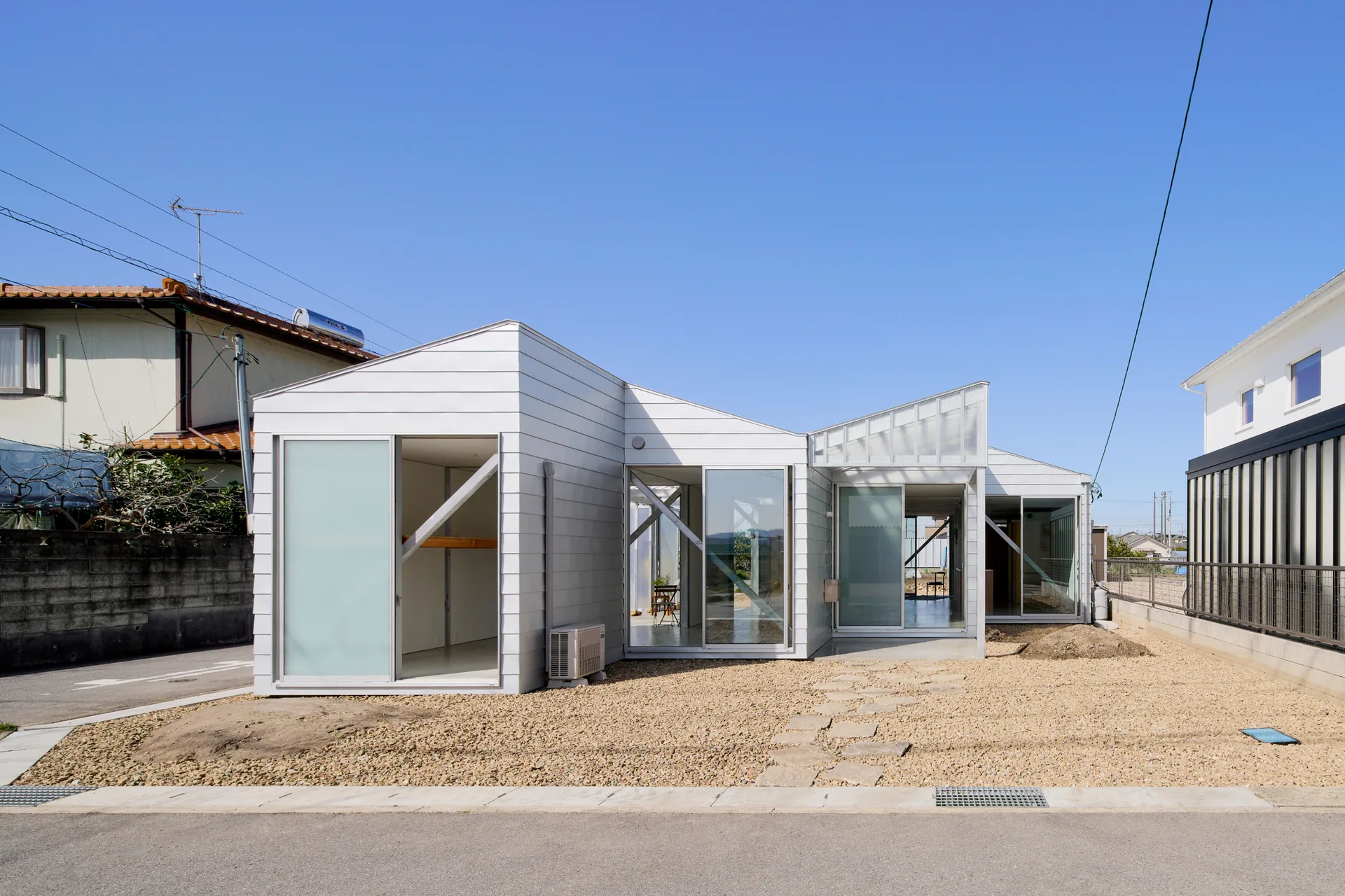 Street view of white clad gabled volumes stepping back from a gravel forecourt under a clear sky, House OY by 1-1 Architects