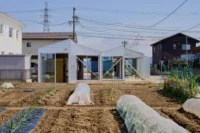 Two white gabled volumes with glazed fronts seen across a vegetable field with plastic tunnel covers, House OY by 1-1 Architects