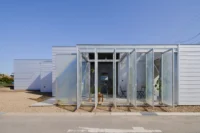 Front of a volume enclosed by tall translucent folding panels within a light frame, behind a gravel strip, House OY by 1-1 Architects