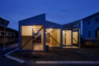 Gabled volumes glowing at dusk through large windows with diagonal braces, seen from the street corner, House OY by 1-1 Architects