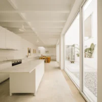 Long kitchen with a white island and joinery beside a full height window onto the courtyard