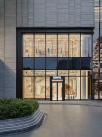 Two storey glazed shopfront lit from inside at dusk, with a curved planter in front, Housing CARNIVAL by Studio Profile