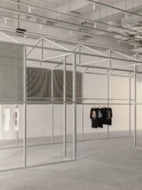 White steel frame structure holding a single rail of printed T shirts in a pale grey room, Housing CARNIVAL by Studio Profile
