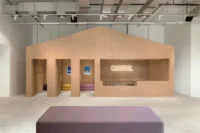 Pale timber facade with a pitched outline, door openings and an illuminated CARNIVAL sign, Housing CARNIVAL by Studio Profile