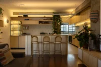 Open kitchen with a high plywood counter, three bar stools and hanging plants under warm cove lighting