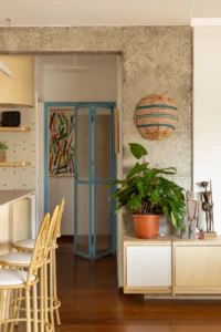 Exposed concrete column next to a blue framed door, a plant on a white cabinet and rattan stools