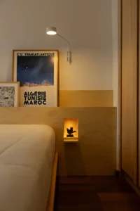 Bedside niche lit within a plywood headboard below a travel poster and wall lamp