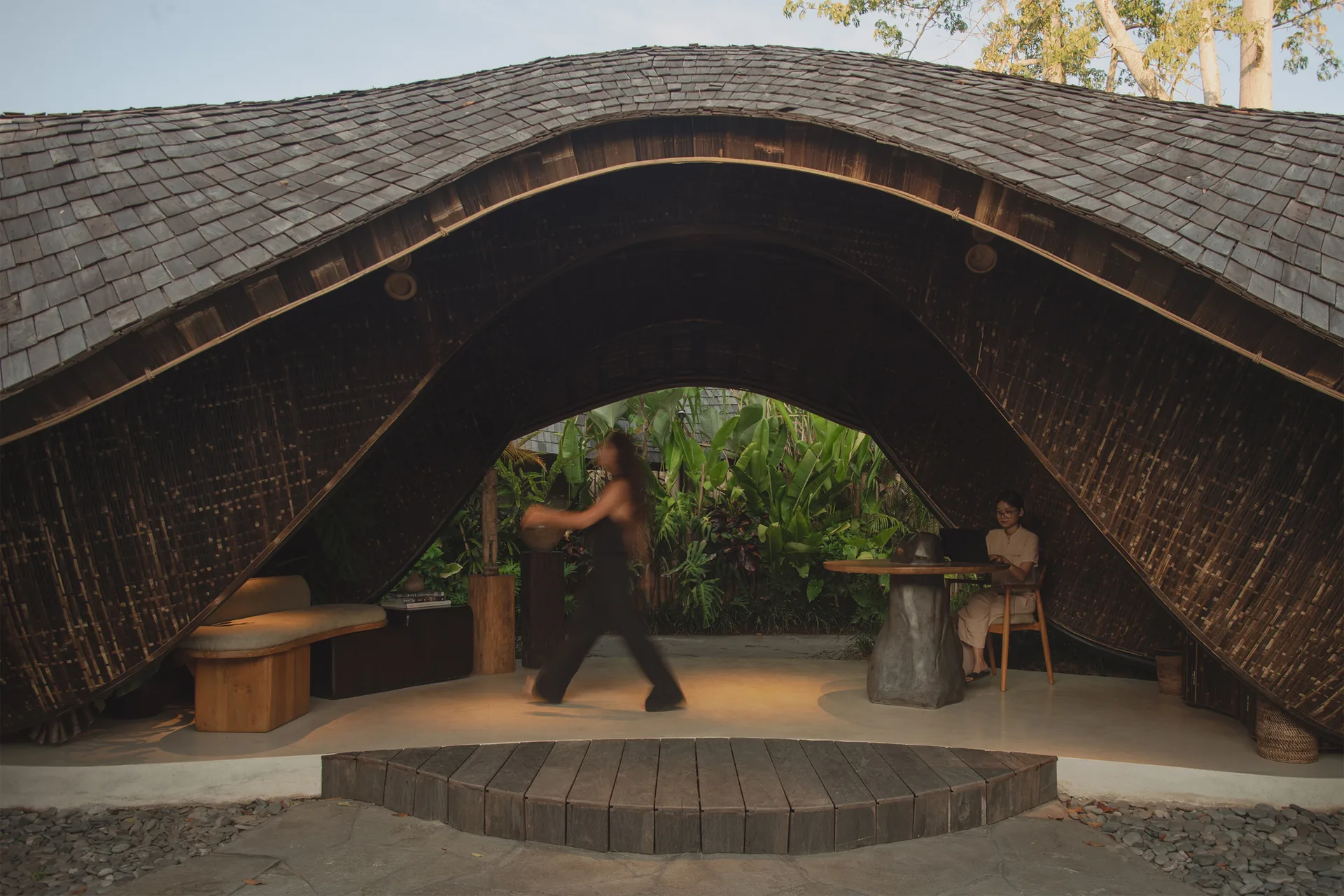 Guest walking beneath the wave shaped roof past a bench and a round reception desk