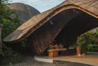 Side view of the curved roof sweeping down to the ground over a cushioned bench in tropical planting