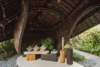 Lounge corner under the ribbed bamboo ceiling with a cushioned bench, plants and carved timber objects