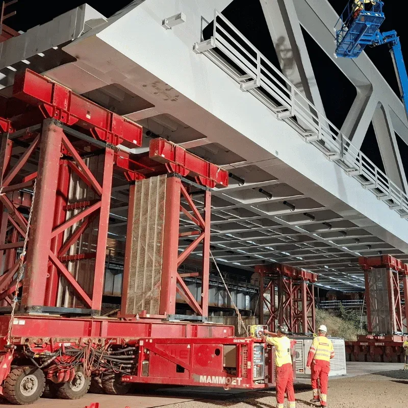 Close view of red support masts on a modular transporter under the white steel deck of a bridge, with workers alongside
