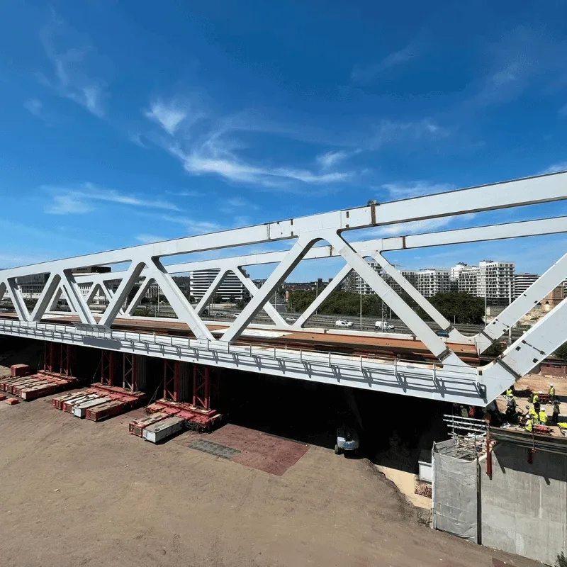 A white steel truss bridge spanning a road on concrete abutments, seen from above with apartment blocks behind