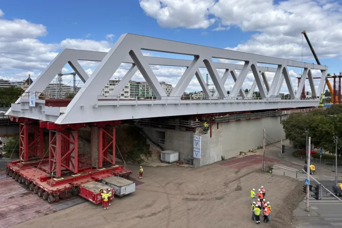 A white steel truss bridge resting on red support towers and modular transporters on a construction site, with workers in hi-vis beside it