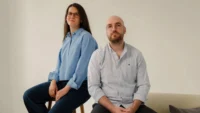 A woman and a man in light blue shirts sitting side by side against a pale wall