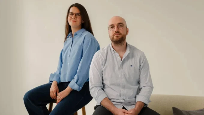 A woman and a man in light blue shirts sitting side by side against a pale wall