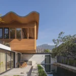 Timber upper volume with a wavy roof edge projecting over a white brick house and a long reflecting pool, Los Navegantes House