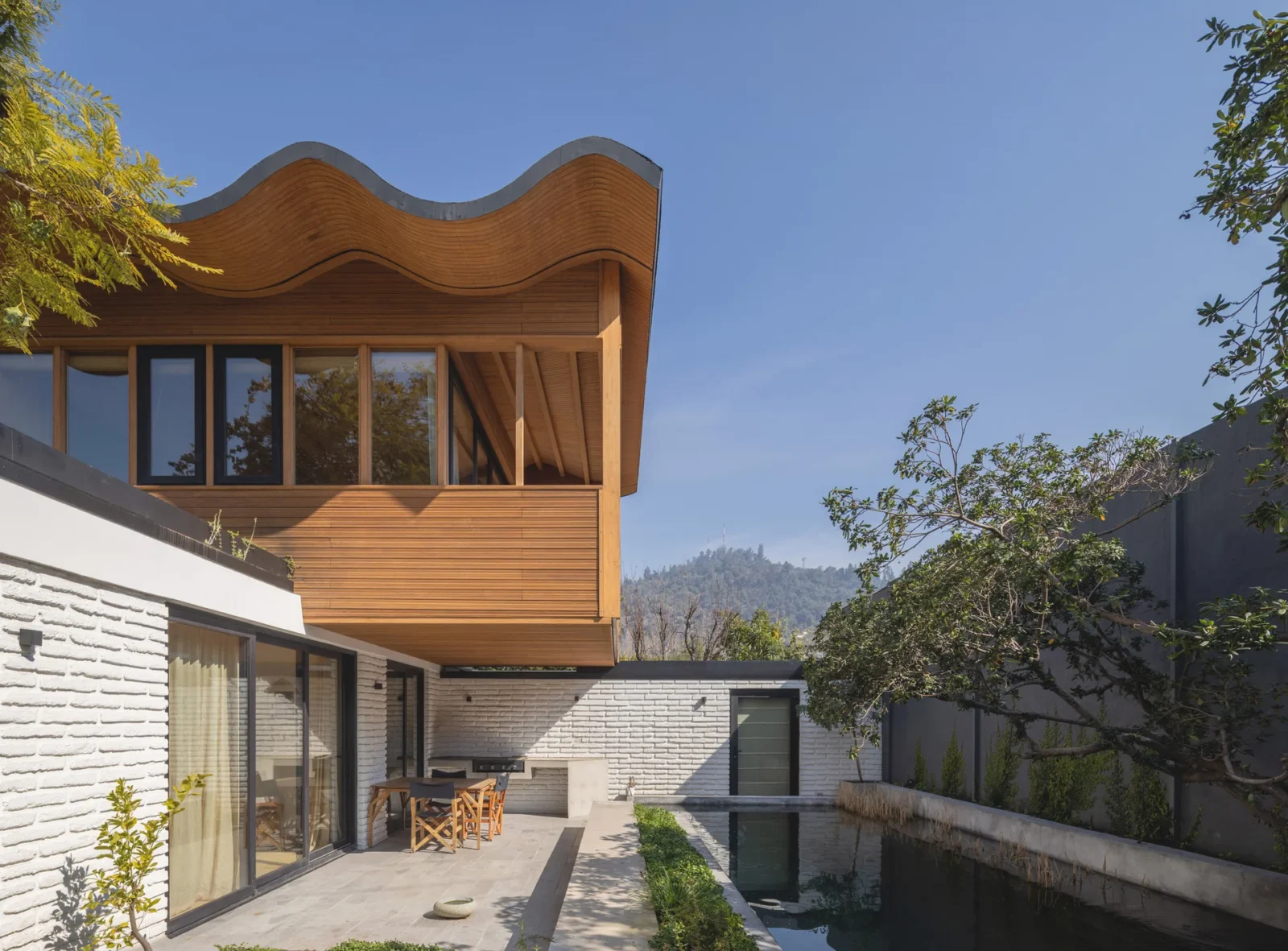 Timber upper volume with a wavy roof edge projecting over a white brick house and a long reflecting pool, Los Navegantes House