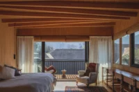 Upstairs bedroom under exposed timber beams with an armchair and a balcony looking toward the hill, Los Navegantes House