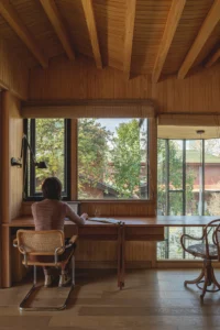 Person working at a desk by a corner window in a room with timber ceiling joists, Los Navegantes House