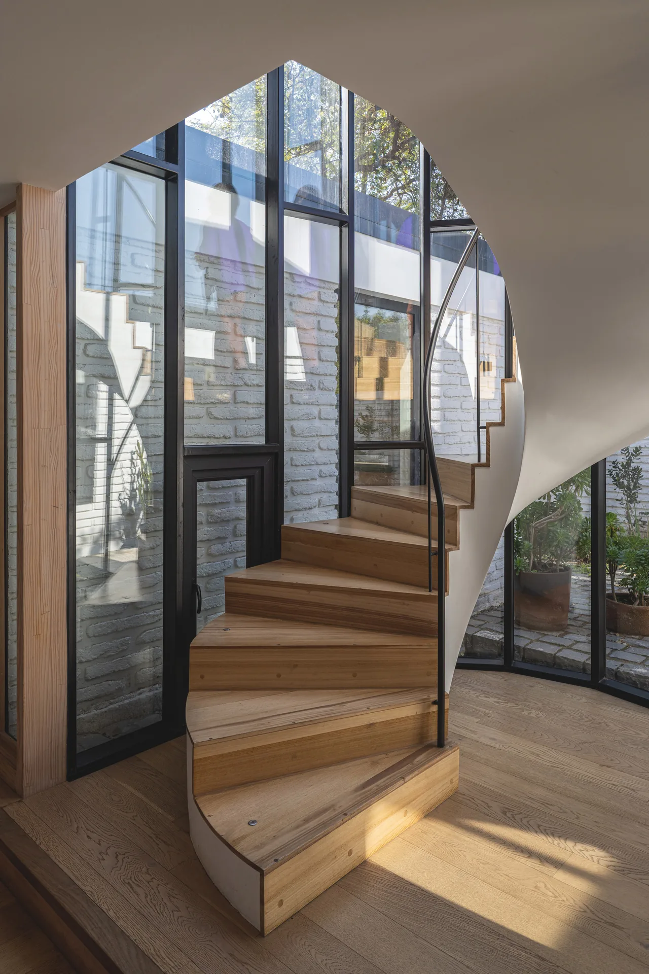 Wooden spiral stair rising inside a curved glass enclosure with the courtyard beyond, Los Navegantes House
