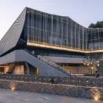 Theater with a pleated facade of vertical steel tubes lit at dusk above a stone retaining wall and plaza, Moganshan Liaoyuan Theater by One Design