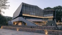 Theater with a pleated facade of vertical steel tubes lit at dusk above a stone retaining wall and plaza, Moganshan Liaoyuan Theater by One Design