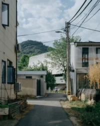 Narrow village lane between houses and utility poles with the theater's pale roof beyond, Moganshan Liaoyuan Theater by One Design