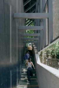 People climbing a narrow outdoor stair between dark walls and concrete frames, Moganshan Liaoyuan Theater by One Design