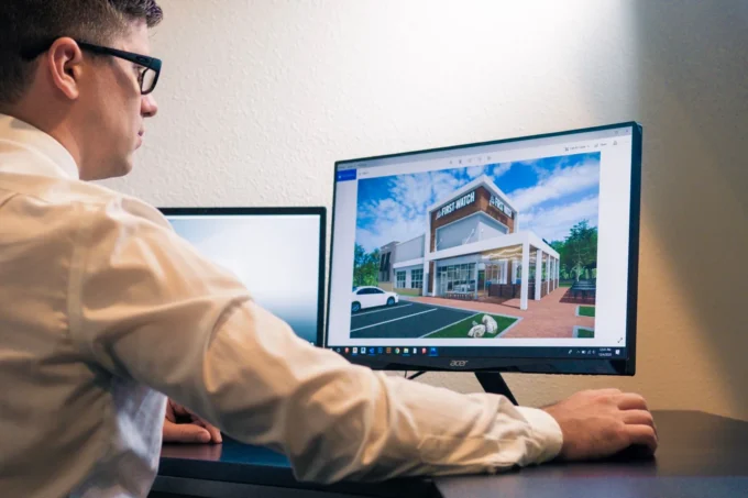 Architect at a desk reviewing a photorealistic exterior render of a house on a large monitor, the kind of image small practices outsource renders for