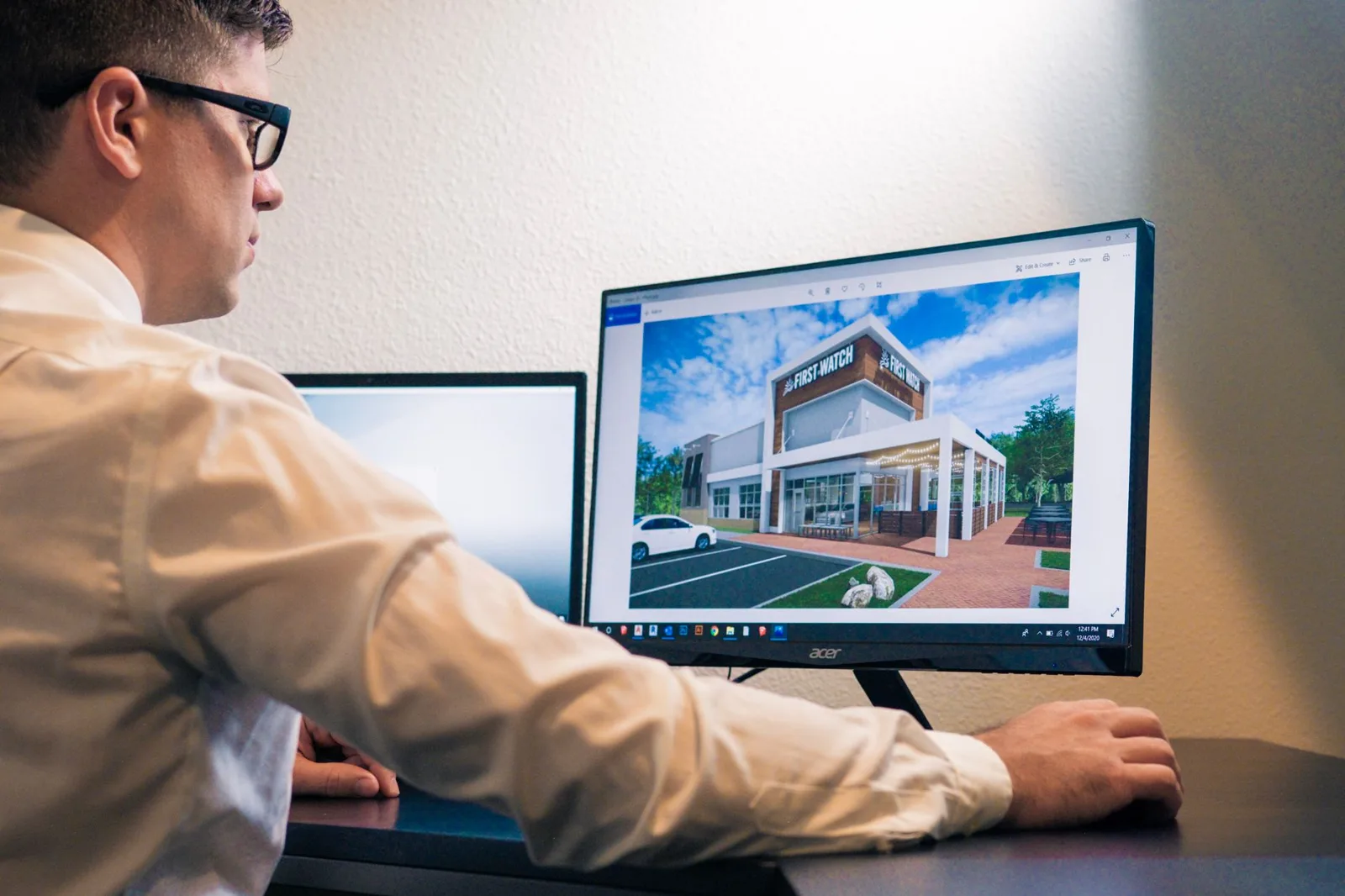 Architect at a desk reviewing a photorealistic exterior render of a house on a large monitor, the kind of image small practices outsource renders for