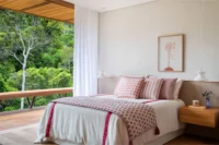 Bedroom with a pink patterned bedspread facing floor to ceiling glass and a timber balcony, Pedra Azul House by Escala Arquitetura