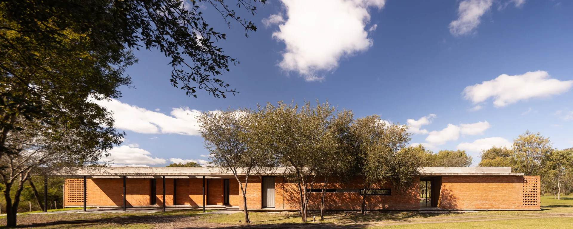 Long horizontal house seen across a meadow between scattered trees under a blue sky